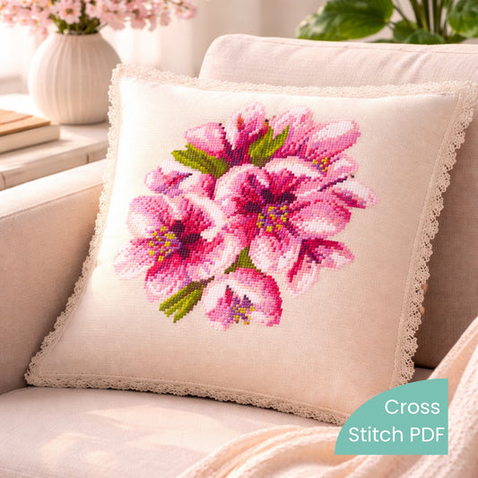 Cherry blossom cross stitch pattern PDF shown on decorative pillow with pink floral embroidery design in cozy living room setting.