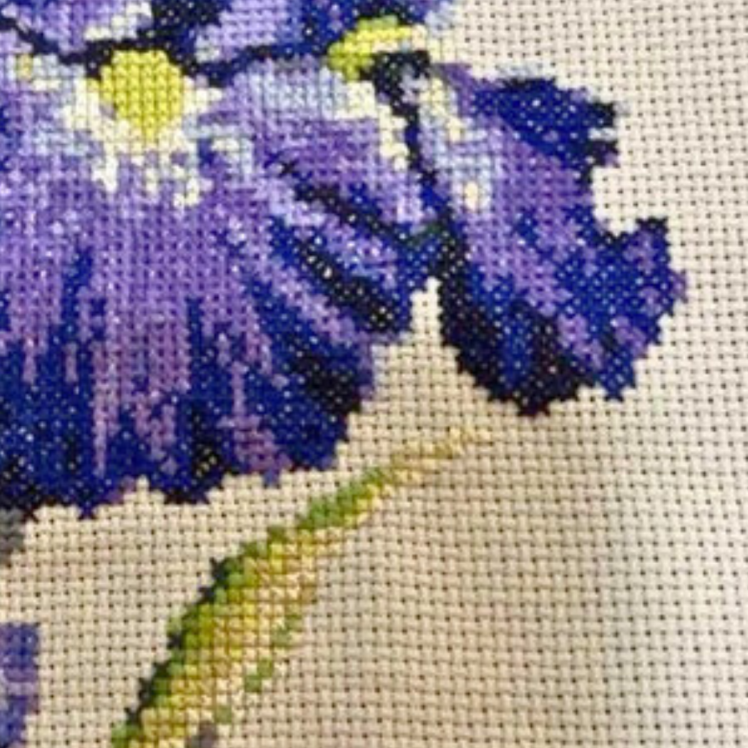 Close-up detail of purple blue iris cross stitch showing smooth color transitions and fine stitching texture on linen fabric