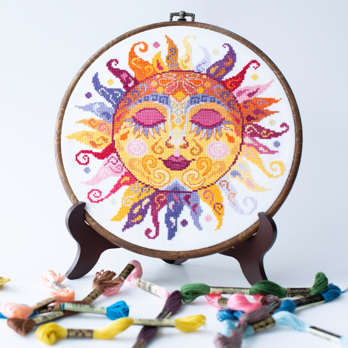 Colorful sun-themed cross-stitch embroidery on a wooden hoop with embroidery floss on a white background.