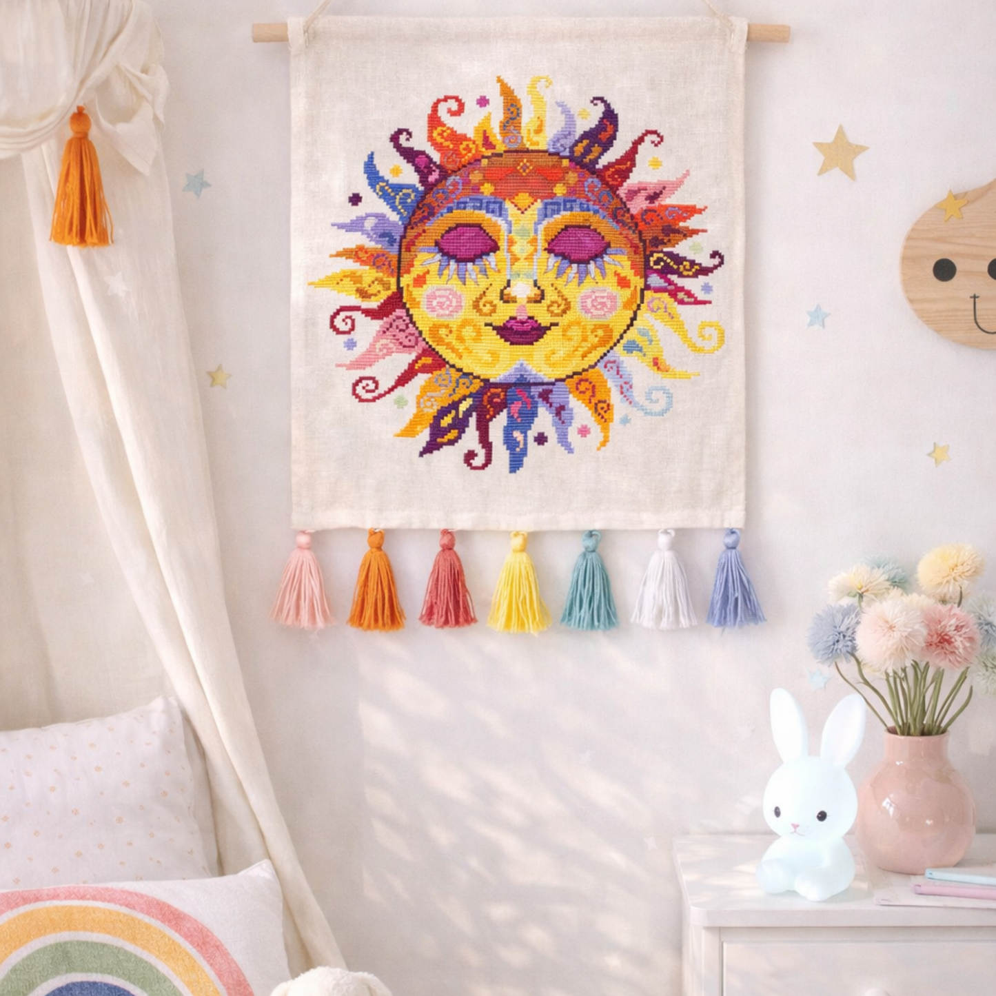 Colorful swirl sun cross stitch pattern used as textile wall hanging with tassels, bright folk embroidery decor for nursery or boho interior