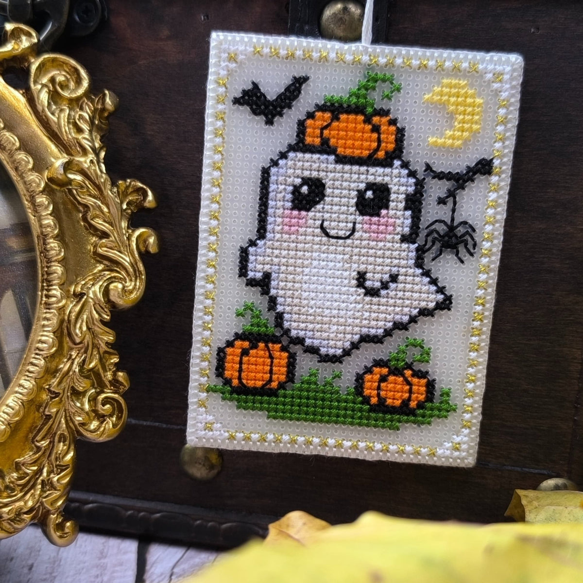 Cross-stitched ghost with pumpkins and a moon on a wooden surface with a gold frame.