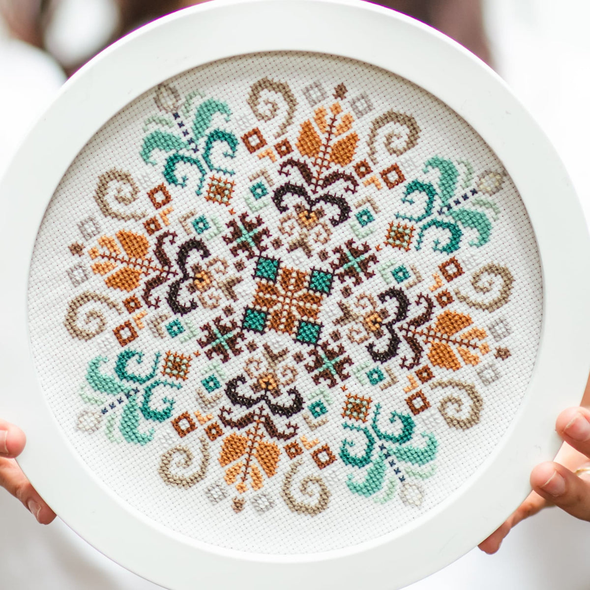 Finished geometric cross stitch embroidery in round hoop, modern folk ornament with symmetrical mandala pattern on white fabric.