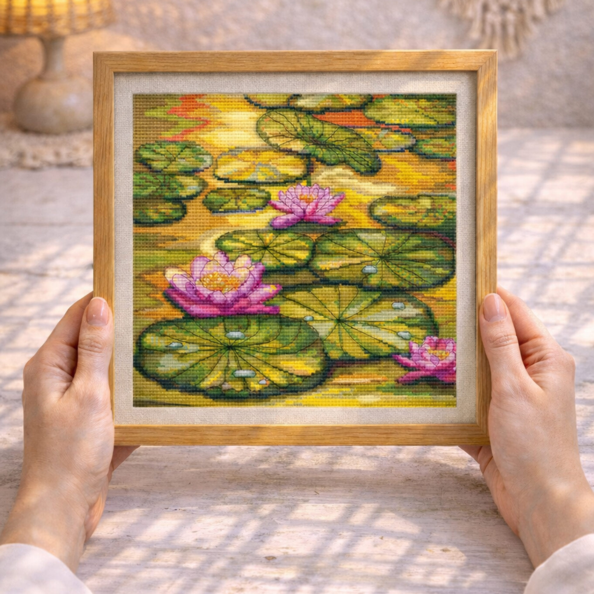 Framed cross-stitched picture of water lilies held by hands on a textured surface.