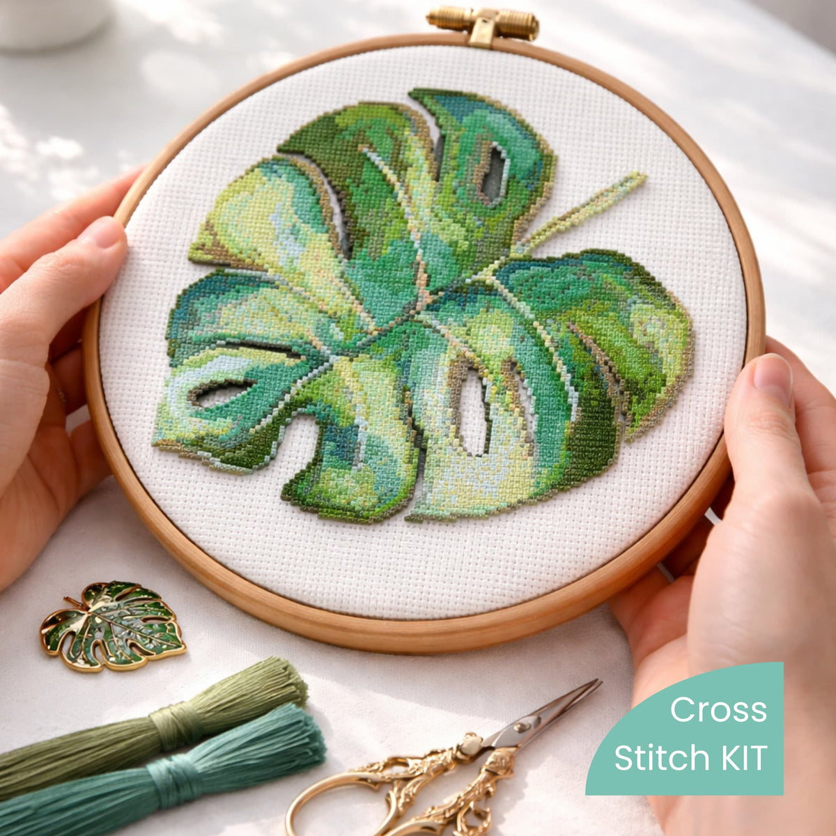 Monstera leaf cross stitch kit displayed in embroidery hoop held in hands, detailed tropical botanical design.