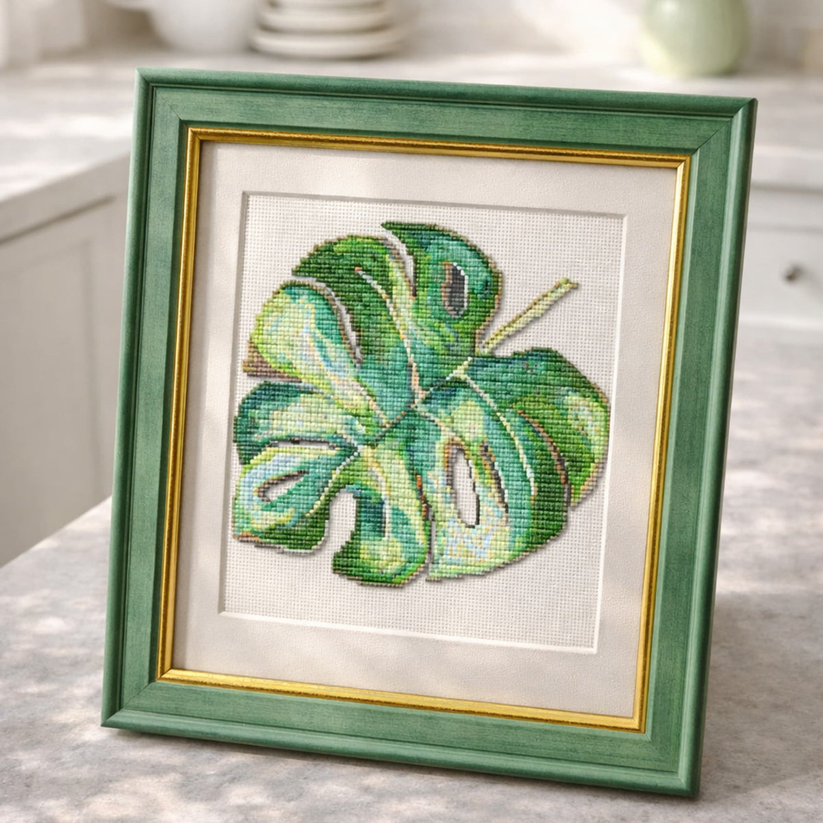 Monstera leaf cross stitch kit shown in green frame as finished botanical embroidery artwork.