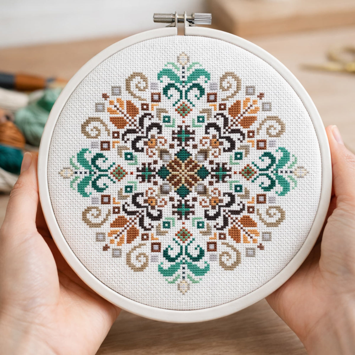 Round hoop cross stitch kit with symmetrical geometric folk motif in earthy and teal colors, held in hands, finished embroidery example.
