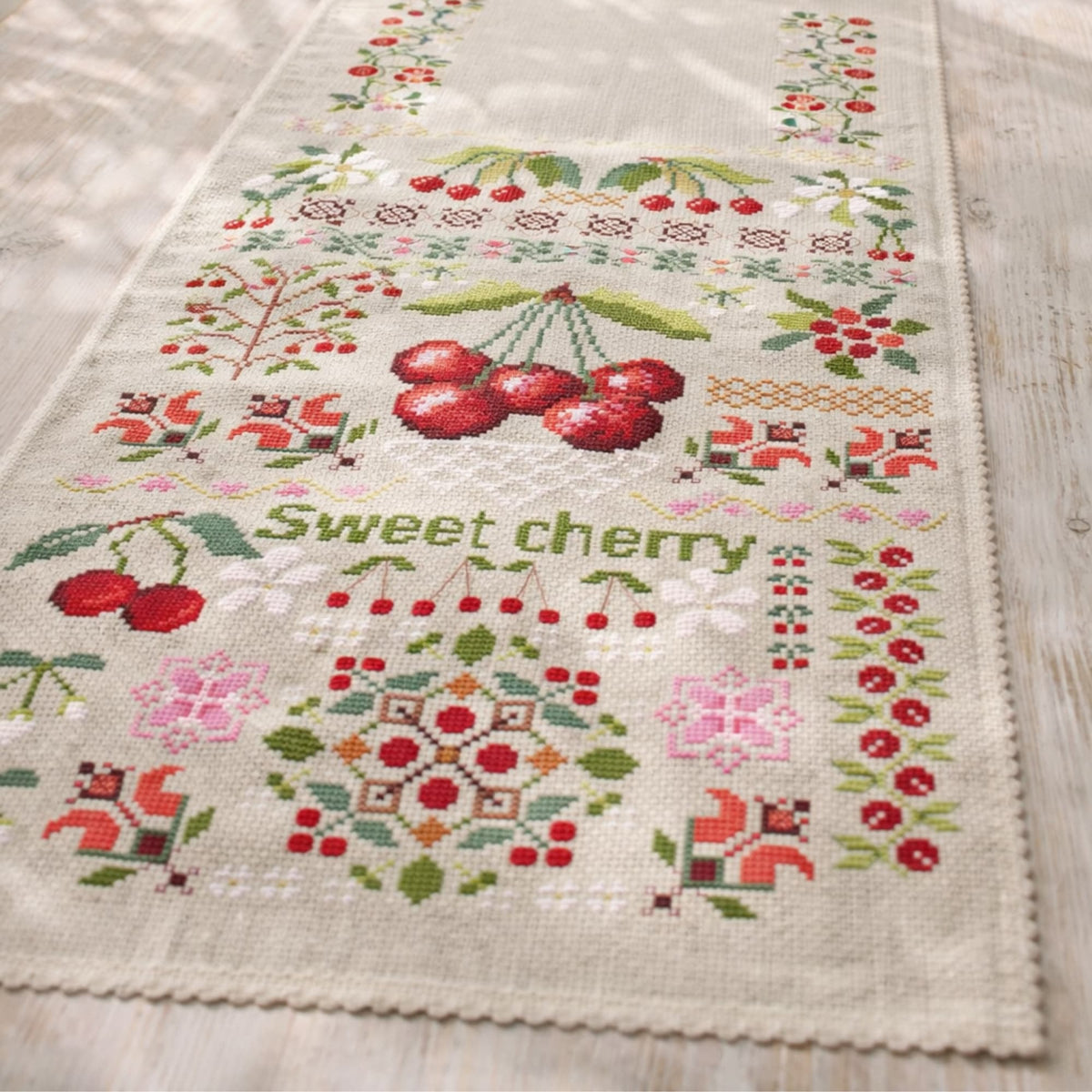 Sweet cherry cross stitch kit shown on fabric with detailed fruit sampler and floral embroidery motifs.